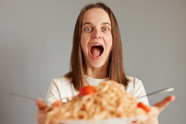 검은 머리에 맛있는 파스타를 먹고 회색 배경 위에 고립된 저녁 식사를 하며 카메라를 바라보며 흥분한 행복한 여성의 초상화