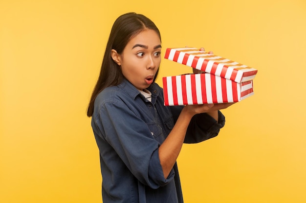 Portrait of amazed funny girl in denim shirt opening gift and looking into box with shocked astonished expression found unbelievable present surprise indoor studio shot isolated on yellow background