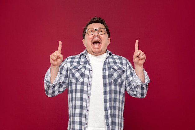 Portrait of amazed funny middle aged man in checkered shirt, eyeglasses standing, looking up, shocked face and pointing at up copyspace on his head. indoor studio shot, isolated on dark red background