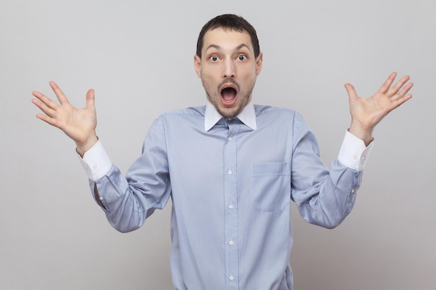 Portrait of amazed handsome bristle businessman in classic blue shirt standing with raised arms and looking at camera with surprised face . indoor studio shot, isolated on grey background copyspace.