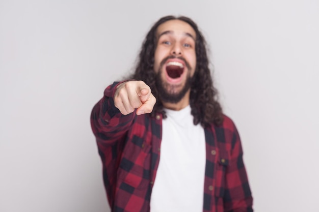 Portrait of amazed handsome man with beard and black long curly hair in casual checkered red shirt standing pointing, looking at camera and surprised. indoor studio shot, isolated on grey background.