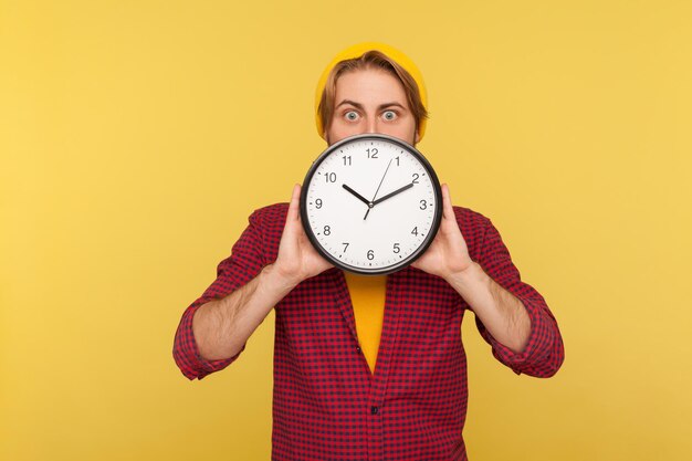 Portrait of amazed hipster guy in checkered shirt hiding face behind big clock and looking at camera with worried shocked expression, showing time to hurry up. studio shot isolated, yellow background