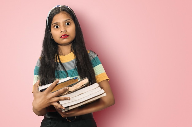 Portrait of a amazed indian asian girl student with open mouth and big eyes posing isolated holding a lot of books and shocked by seeing exam syllabus Education concept