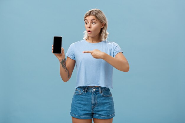 Portrait of amazed shocked attractive blonde girl in trendy summer t-shirt holding smartphone gasping from surprise pointing at gadget screen posing