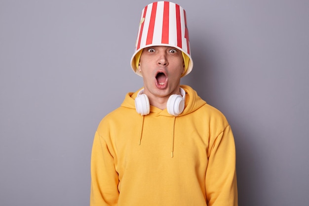 Portrait of amazed shocked emotional hipster man wears yellow hoodie standing against grey wall expressing astonishment posing with empty popcorn bucket on his head screaming watching horror movie