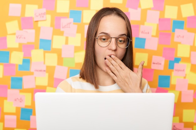 Portrait of amazed shocked surprised woman freelancer with dark hair wearing striped Tshirt posing isolated over yellow background working on laptop covering mouth with palm