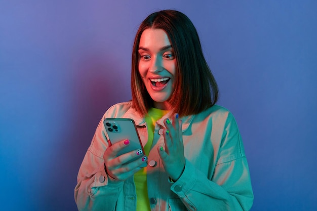 Portrait of amazed surprised Caucasian woman wearing stylish jacket posing isolated on neon light background using cell phone reading breaking news looking at display with big eyes