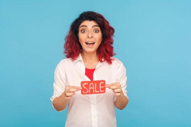 Portrait of amazed trendy woman with fancy red hair holding Sale word and looking at camera with shock and excitement shopaholic surprised by discounts low prices studio shot blue background