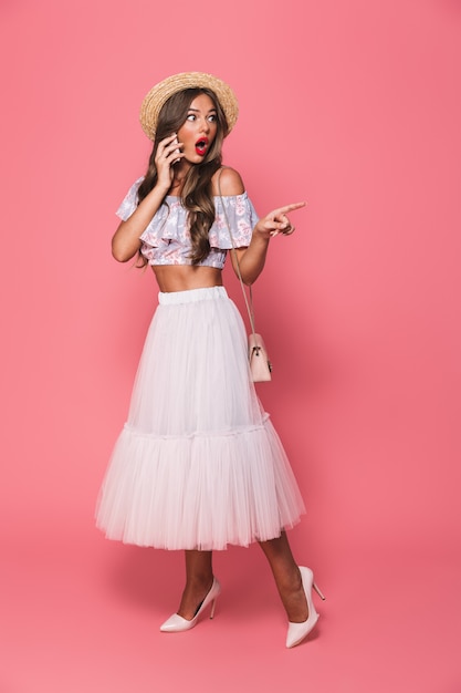 Portrait of amazed woman wearing straw hat and fluffy skirt smiling and talking on cell phone