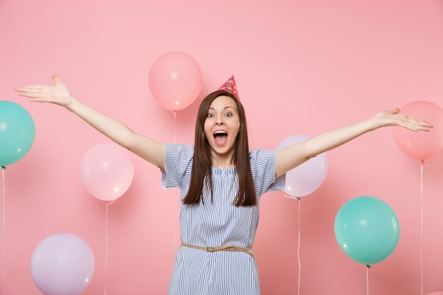 Portrait of amazed young happy woman in birthday hat blue dress screming spreading hands on pastel pink background with colorful air baloons. Birthday holiday party, people sincere emotions concept.