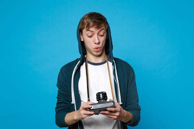 Portrait of amazed young man in casual clothes holding, looking on retro vintage photo camera isolated on blue wall . People sincere emotions lifestyle concept. 