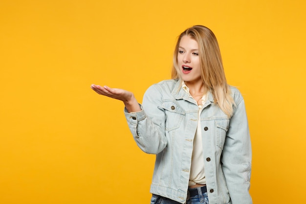 Portrait of amazed young woman in denim casual clothes pointing hand aside, holding something isolated on bright yellow orange wall background in studio. People lifestyle concept. Mock up copy space.