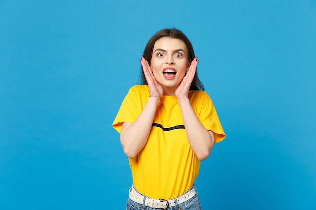 Portrait of amazed young woman in vivid casual clothes looking camera, keeping mouth open, spreading hands isolated on bright blue background in studio. People lifestyle concept. Mock up copy space.