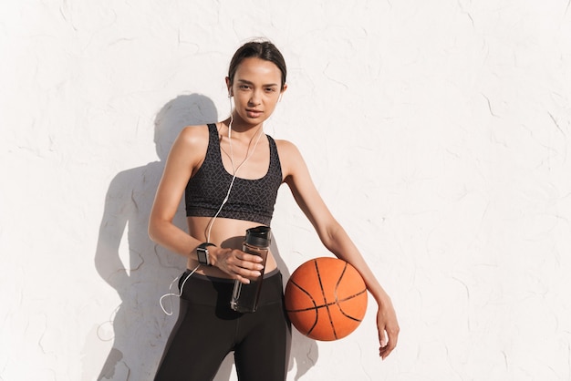 Portrait of amazing concentrated strong sports woman outdoors over white wall posing listening music with earphones holding ball and bottle with water.