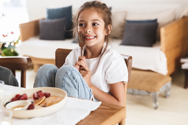 Photo portrait of amazing little girl smiling while having breakfast and eating pancakes with raspberry in bright kitchen at home