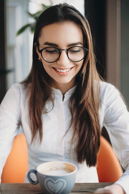 Portrait of a amazing long haired woman wearing eye glasses looking down smiling while sitting in a coffee shop drinking coffee while traveling.