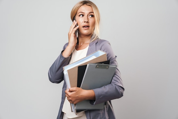 Portrait of an amazing shocked young pretty woman posing isolated over grey wall holding folders talking by mobile phone