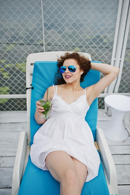 Portrait of an amazing young girl wearing sunglasses enjoying her cocktail sitting on a lounger in lakeside