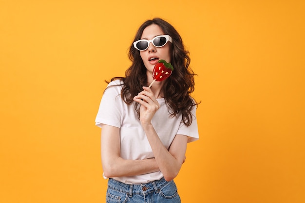 Portrait of amazing young woman posing isolated over yellow wall holding candy lollipop