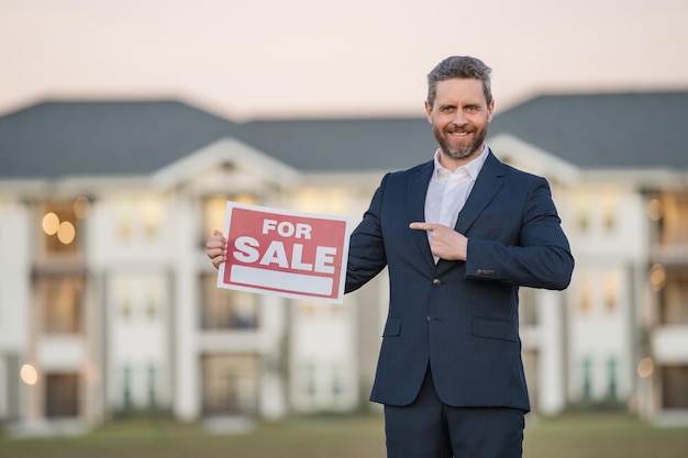 Photo portrait of american real estate agent standing outdoors at house with board for sale real estate