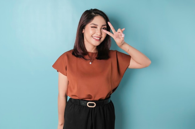 Portrait of amusing Asian woman in casual tshirt holding two fingers or peace sign isolated over blue background