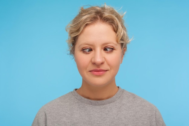 Portrait of amusing comic woman with short curly hair making silly face with crossed eyes, awkward stupid expression, fooling around, having fun. indoor studio shot isolated on blue background