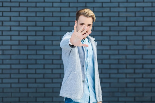 Portrait of anger handsome young blonde man in casual style standing with stop sign gesture and looking at camera focus on hand indoor studio shot on brick wall background