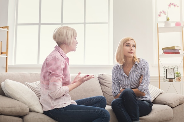 Portrait of angry adult mother shouting and swearing daughter while young woman sitting and listening. Generation and relationship concept