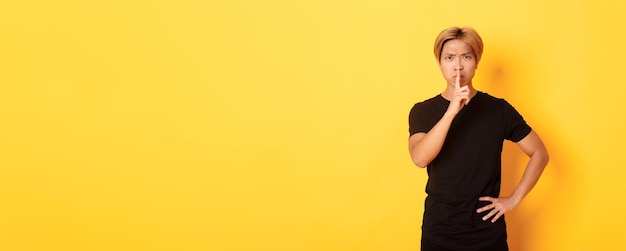 Portrait of angry asian man frowning shushing at camera disappointed standing yellow background