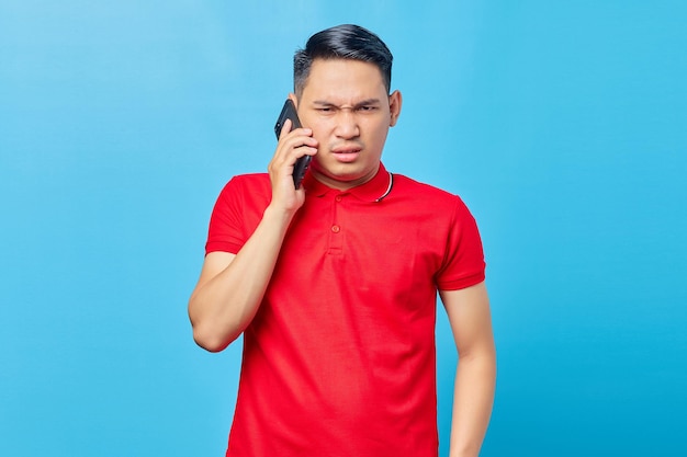 Portrait of angry asian man talking on smartphone and looking at camera isolated on blue background