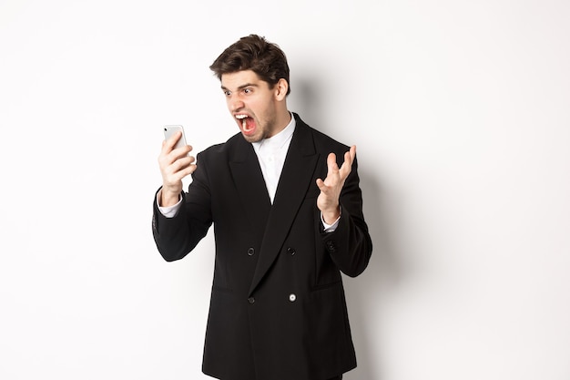 携帯電話で叫んで、ビデオ通話で議論を持ち、白い背景の上に怒って立っている黒いスーツを着た怒っているビジネスマンの肖像画。