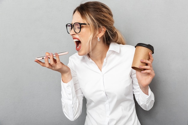 Portrait of angry businesswoman wearing glasses screaming on cell phone and holding coffee in the office, isolated 