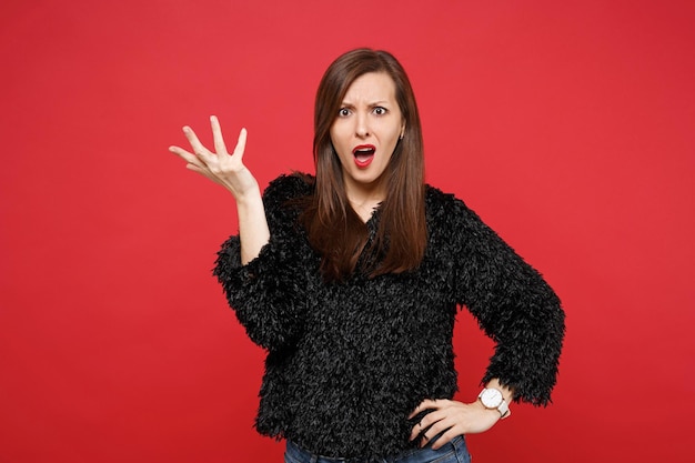 Portrait of angry disgusted young woman in black fur sweater spreading hand, swearing isolated on bright red wall background in studio. People sincere emotions, lifestyle concept. Mock up copy space.