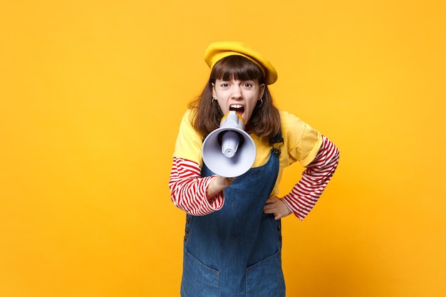 Photo portrait of angry girl teenager in french beret, denim sundress scream in megaphone, swearing isolated on yellow background in studio. people sincere emotions, lifestyle concept. mock up copy space.