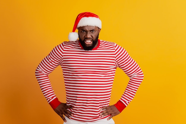 Portrait of angry guy in santa hat hands hips isolated on yellow background