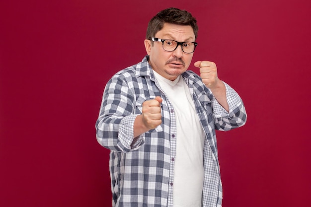 Photo portrait of angry handsome middle aged business man in casual checkered shirt and eyeglasses standing with boxing fists and ready to attack. indoor studio shot, isolated on dark red background.