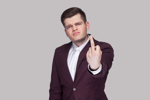 Portrait of angry handsome young man in violet suit and white shirt, standing, looking at camera and showing middle finger. focus on hand. indoor studio shot, isolated on grey background.