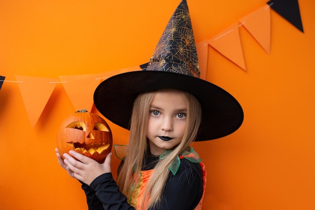 Photo portrait of an angry little girl with a pumpkin in a witch costume on an orange halloween background