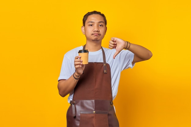 Portrait of angry man wearing apron holding paper cup and showing refusal with thumbs down gesture