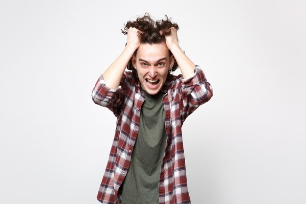 Portrait of angry screaming young man in casual clothes looking camera putting hands on head isolated on white wall background in studio. People sincere emotions lifestyle concept. Mock up copy space.