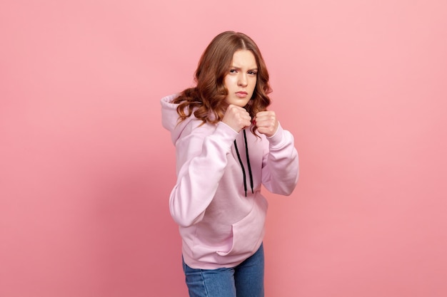 Portrait of angry teen female in hoodie standing with clenched fists and furious look, ready to punch. Indoor studio shot, isolated on pink background