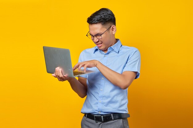 Portrait of angry young asian man Asian in glasses using laptop isolated on yellow background businessman and entrepreneur concept