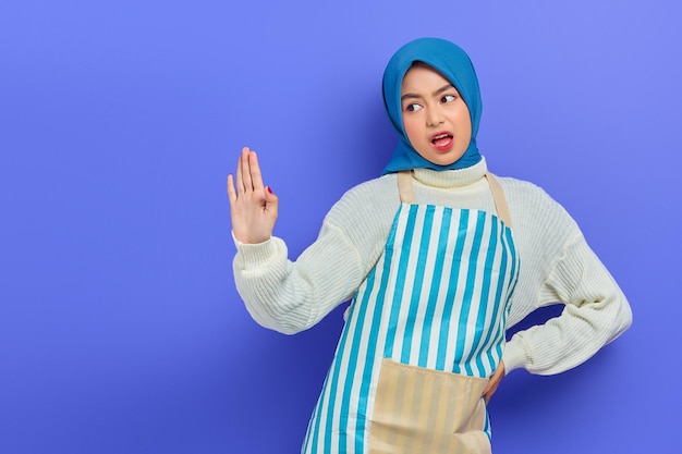 Portrait of angry young asian muslim woman in hijab and striped\
apron showing stop gesture with palm refusing say no isolated on\
purple background people housewife muslim lifestyle concept