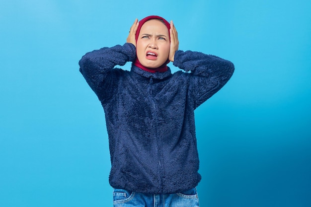 Portrait of angry young Asian woman covering her ears with hand on blue background