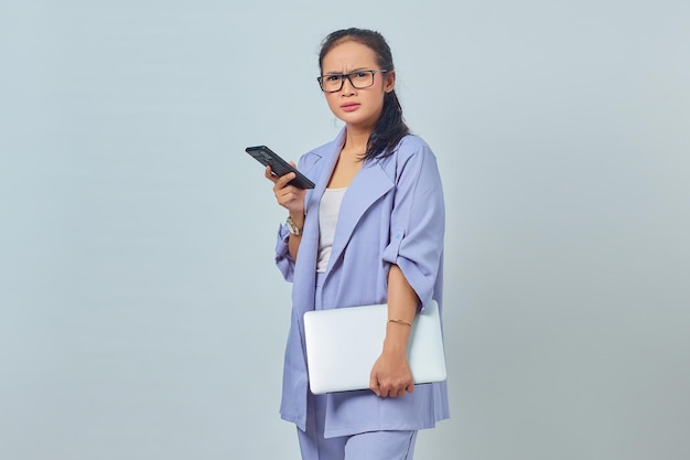 노트북을 들고 흰색 배경에 격리된 휴대전화를 사용하는 화난 젊은 아시아 여성의 초상화