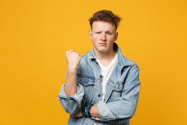 Portrait of angry young man in denim casual clothes looking camera, showing clenching fist isolated on yellow orange wall background. People sincere emotions, lifestyle concept. Mock up copy space.