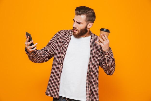 Ritratto di un giovane arrabbiato che indossa abiti casual parlando al telefono cellulare bere caffè da asporto