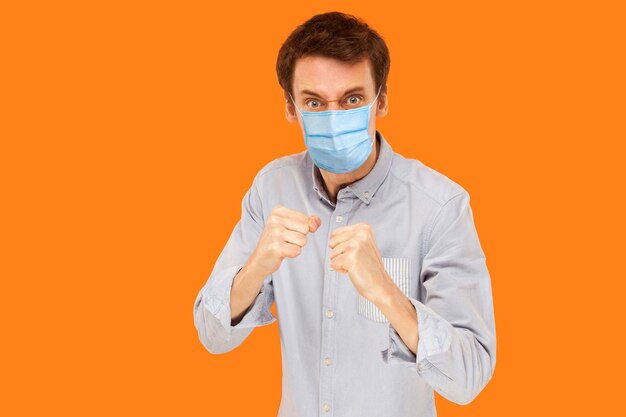 Portrait of angry young worker man with surgical medical mask standing in boxing fists and looking at camera and ready to attack against virus. indoor studio shot isolated on orange background