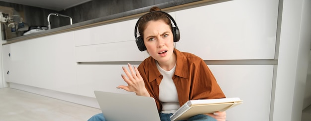 Photo portrait of annoyed and confused woman in headphones sits on floor with laptop and notebook looks