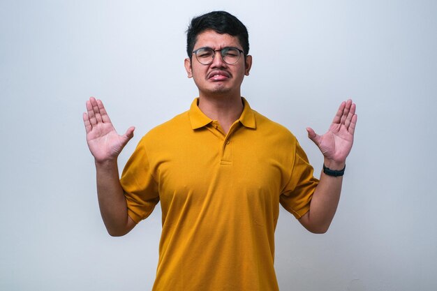 Портрет раздраженного разочарованного азиата, стоящего с поднятыми руками и спрашивающего, почему крытая студия снималась на белом фоне